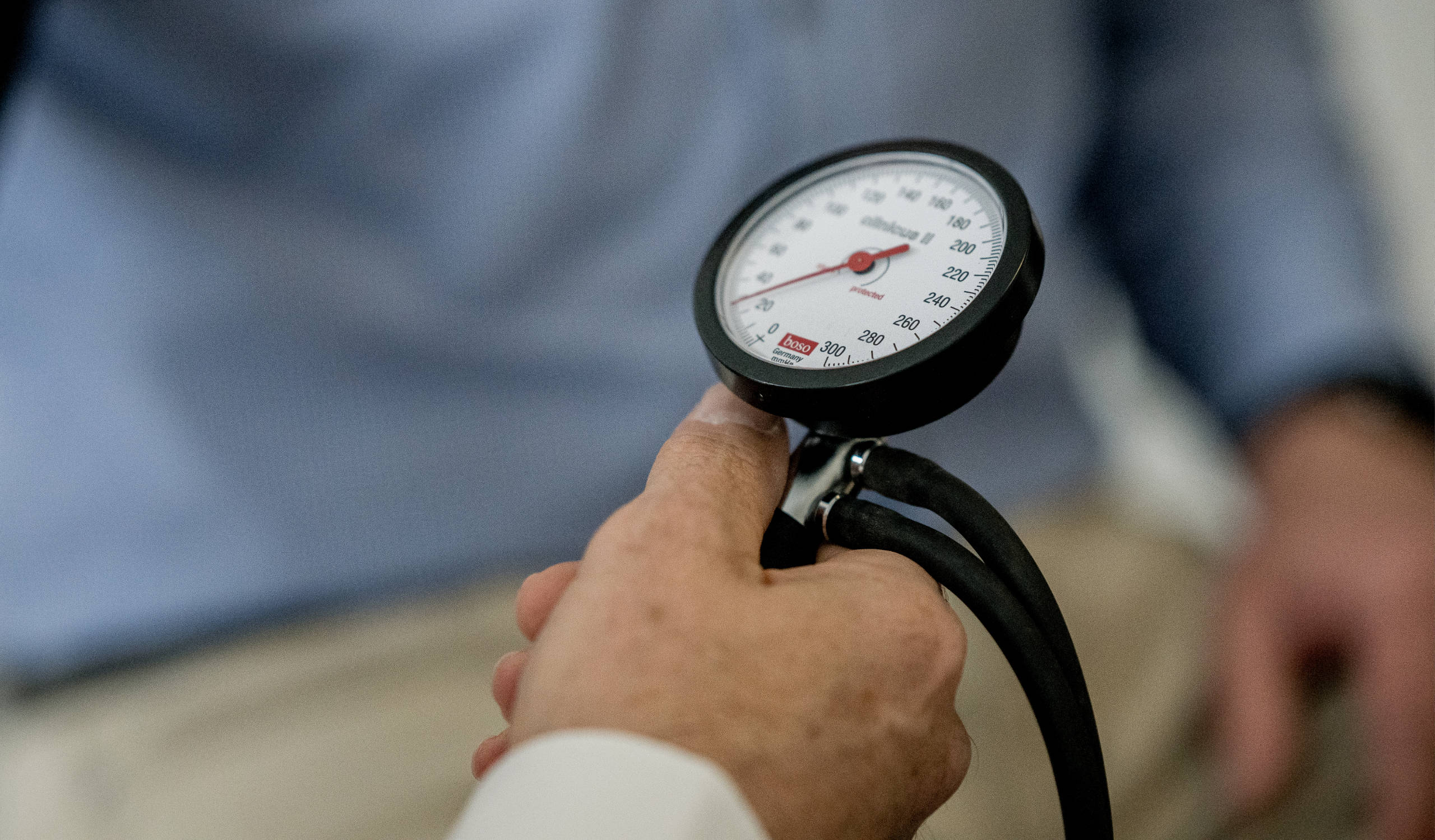 Hand hält Blutdruckmessgerät mit Manometer, unscharf im Hintergrund sitzende Person in blauem Hemd