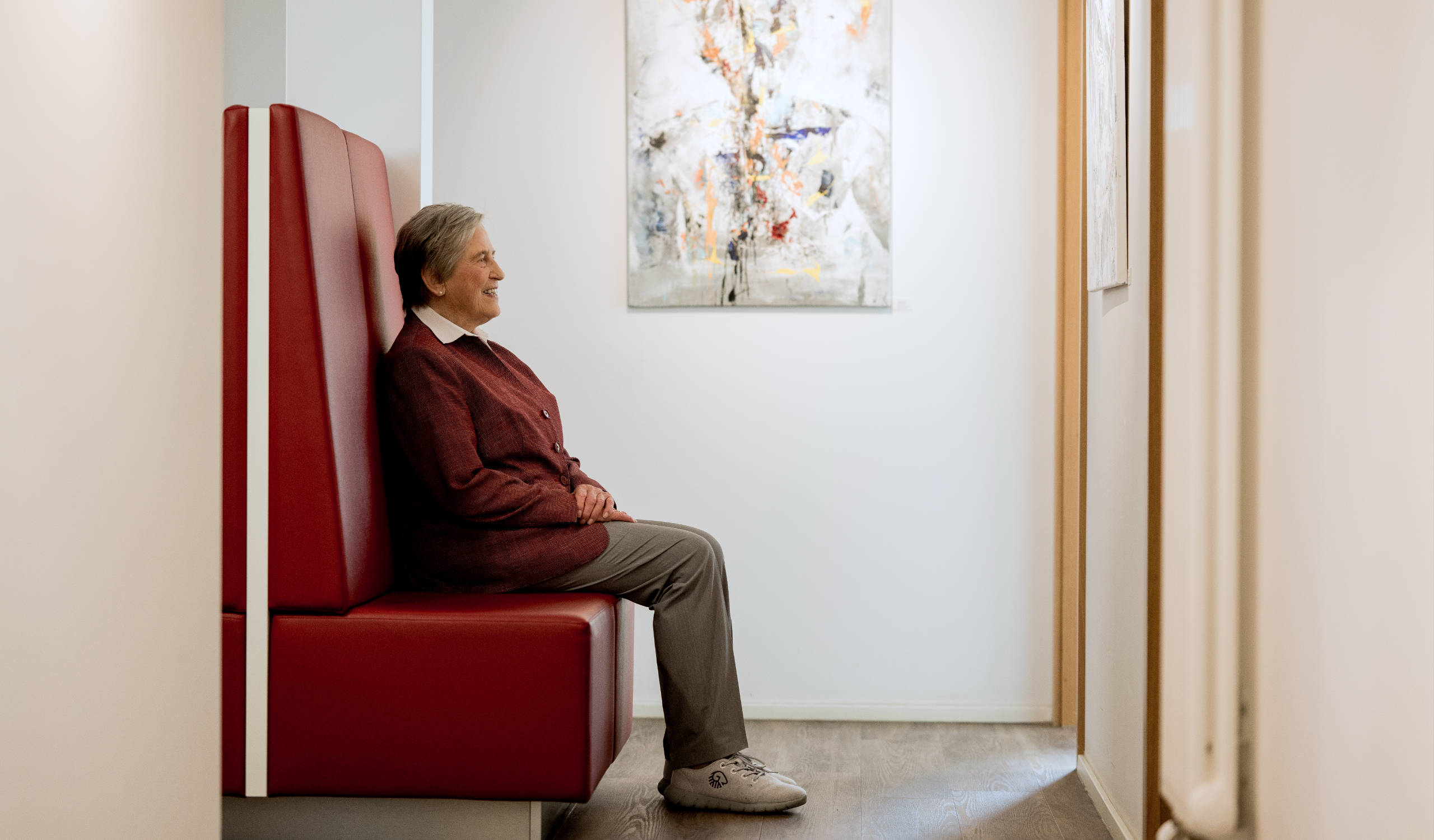 Ältere Frau sitzt wartend auf rotem Sitz in hellem Flur mit abstraktem Bild an der Wand