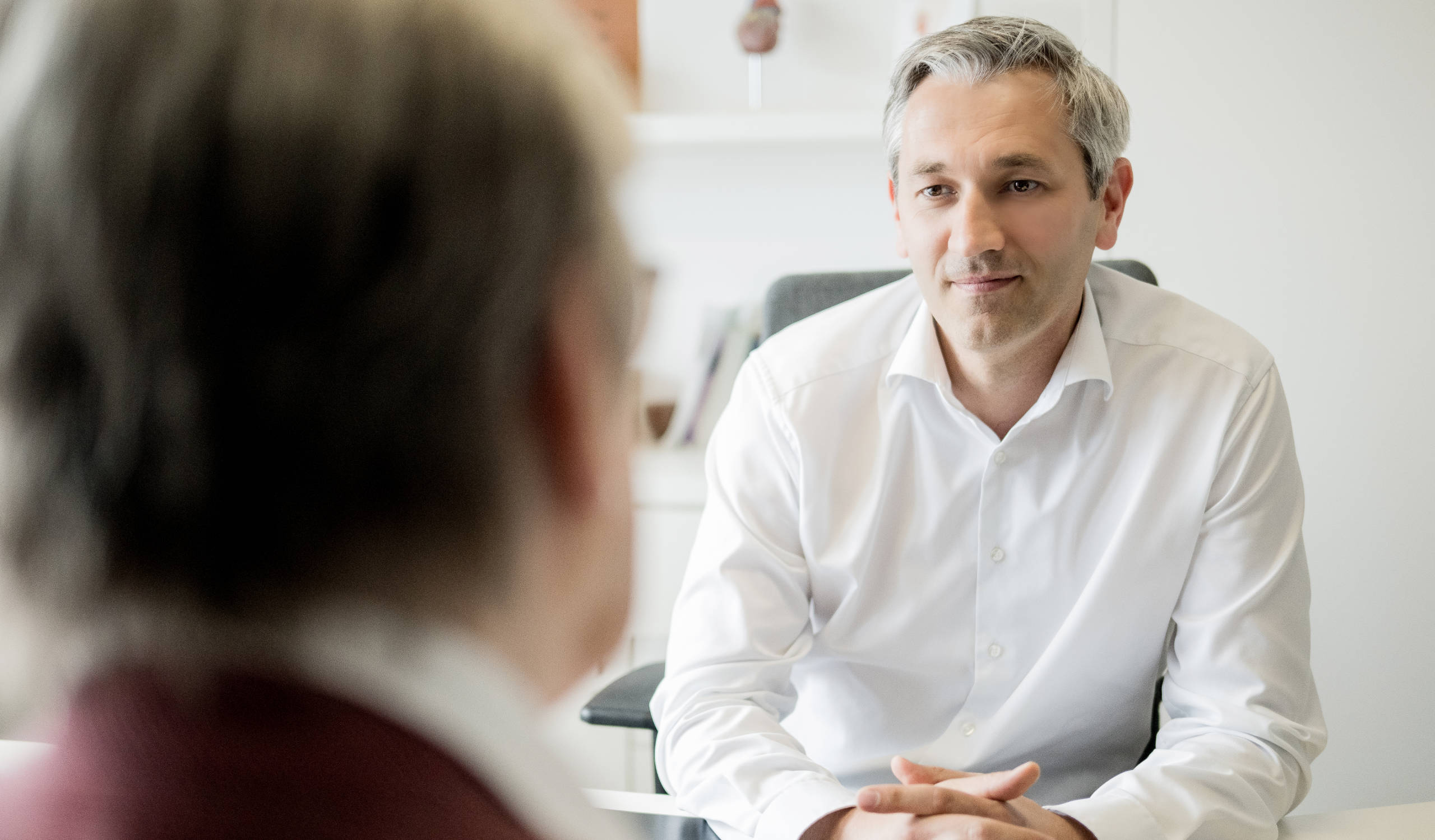 Arzt und Patientin im Gespräch, der Arzt trägt ein weißes Hemd und sitzt mit verschränkten Händen am Tisch