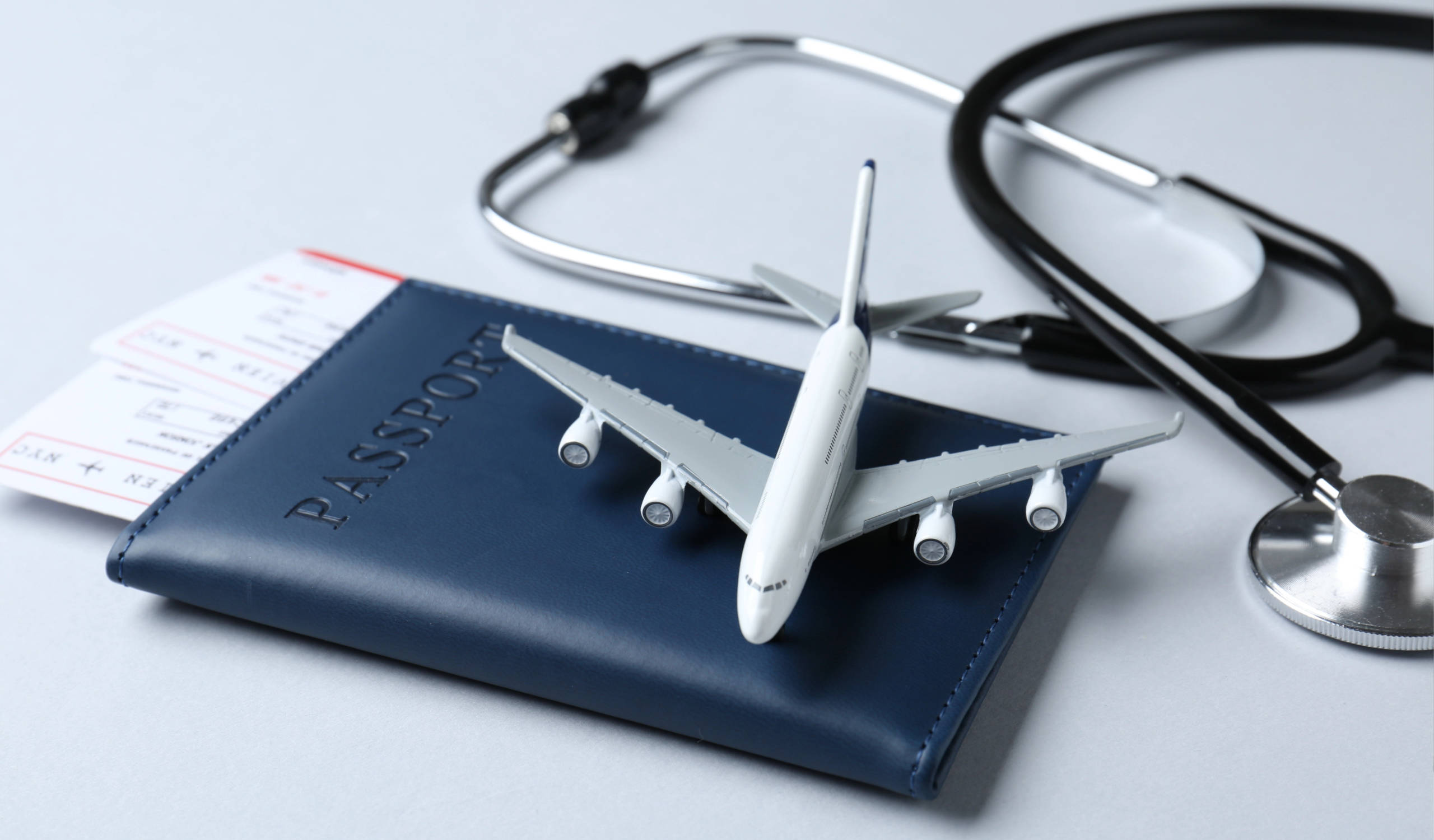 Modellflugzeug auf blauem Reisepass mit Flugticket und Stethoskop im Hintergrund