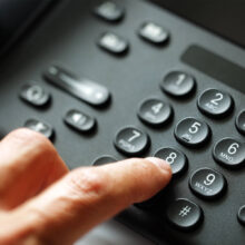 Finger drückt Taste auf einem schwarzen Festnetztelefon mit Zifferntasten